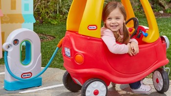 A smiling child sits in a red toy car next to a play electric pump in a grassy outdoor setting.