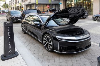 A black electric vehicle with its front trunk open is parked on a city street next to a Lucid Motors sign.