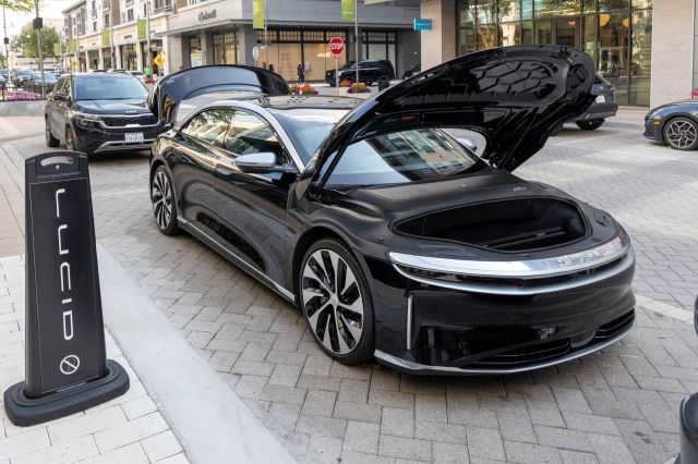A black electric vehicle with its front trunk open is parked on a city street next to a Lucid Motors sign.