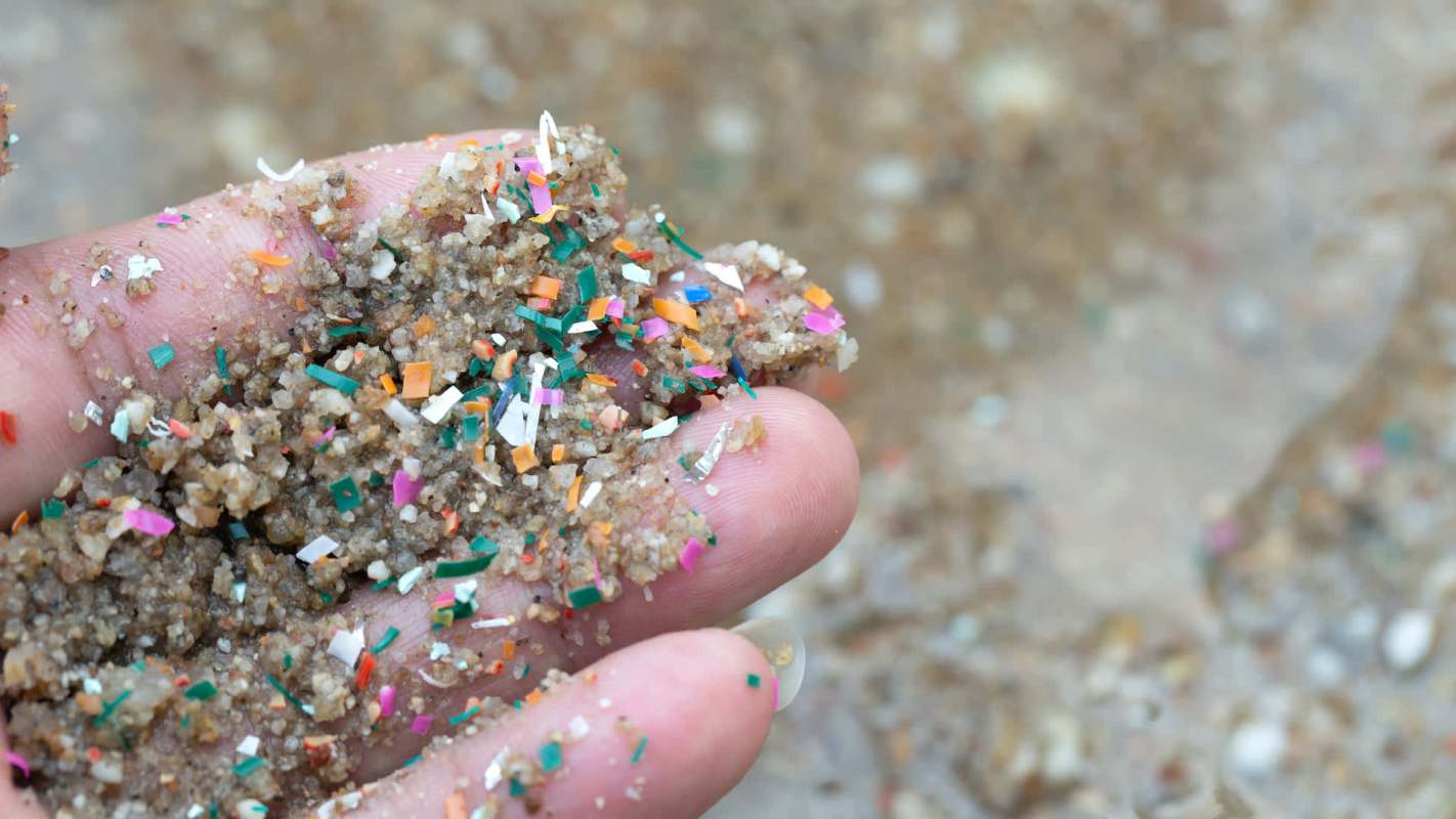 A hand holds a mixture of sand and colorful plastic debris on a blurred sandy background.