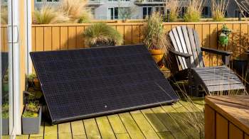 A solar panel rests on a wooden deck next to potted plants and an Adirondack chair in a sunny outdoor space.