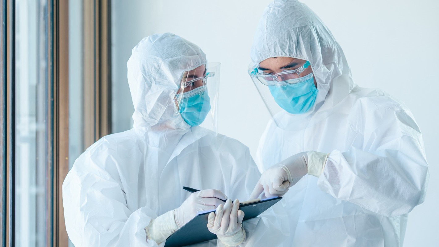 Two healthcare workers in protective gear discuss notes while reviewing data on a clipboard.