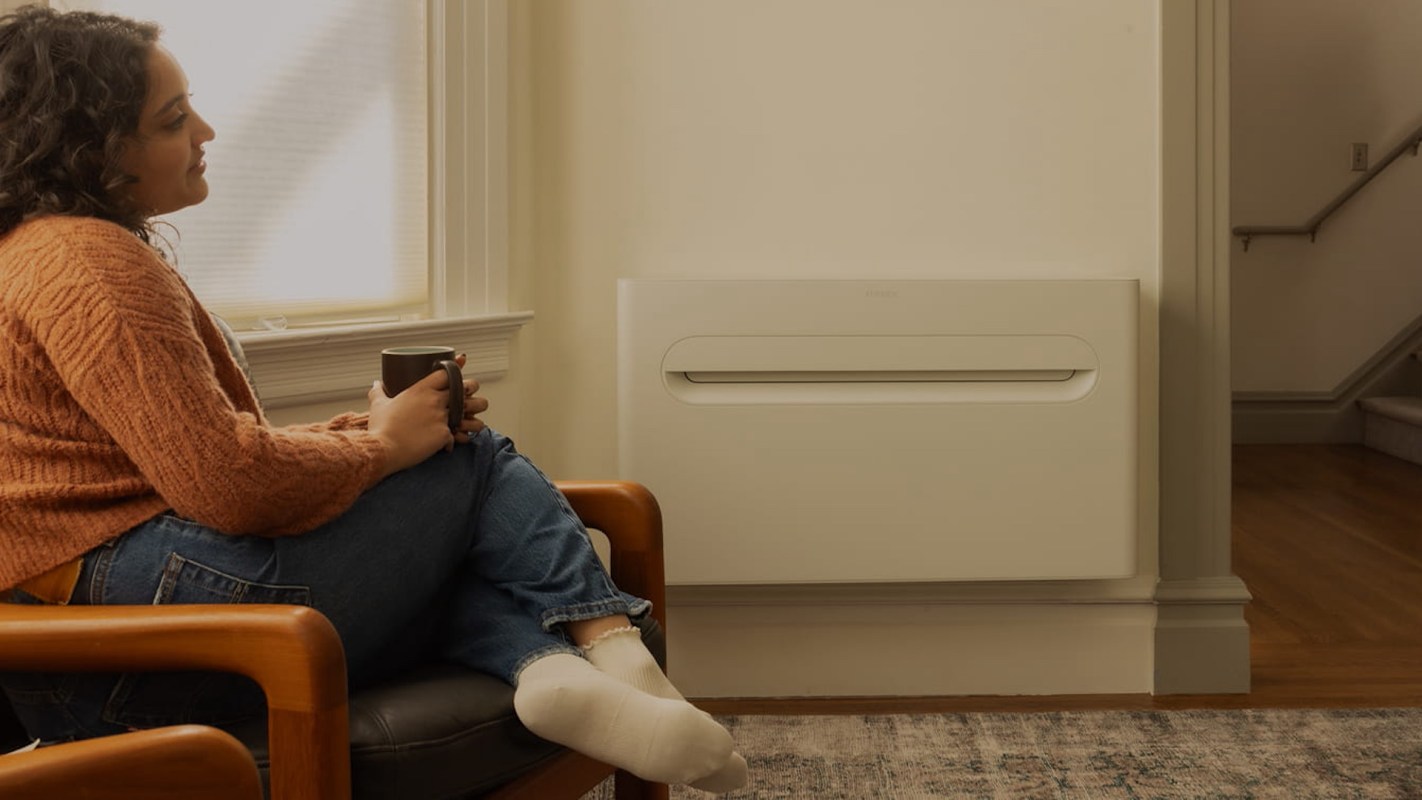 A woman in an orange sweater sits on a chair, holding a coffee mug near a wall-mounted heater.