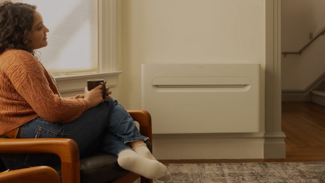 A woman in an orange sweater sits on a chair, holding a coffee mug near a wall-mounted heater.