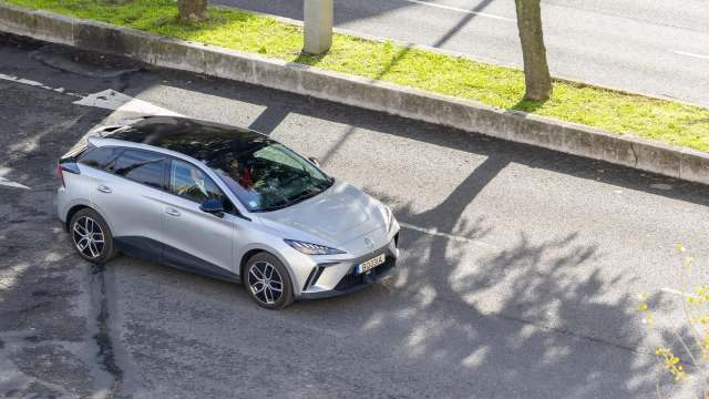 A silver electric car parked on a tree-lined street, with shadows cast on the asphalt.