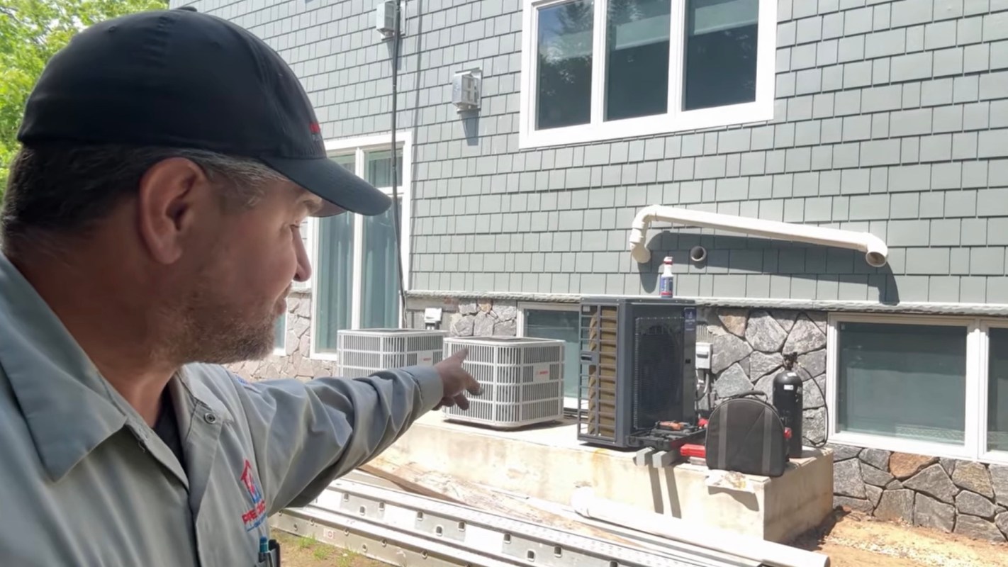 A technician points towards outdoor HVAC units installed beside a house with a stone base and green siding.
