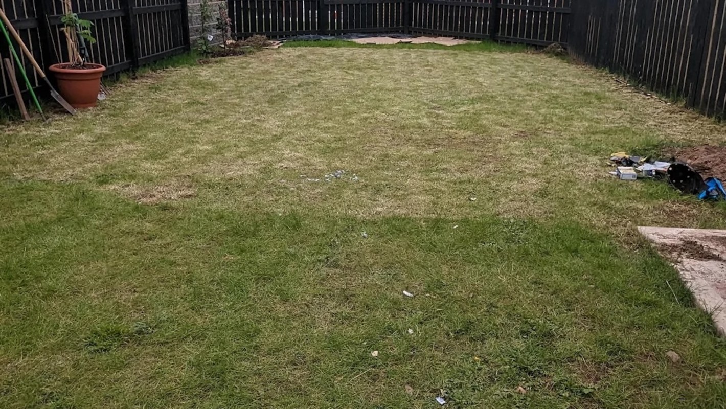 A spacious backyard with uneven grass, a flowerpot, and scattered garden tools against a black wooden fence.