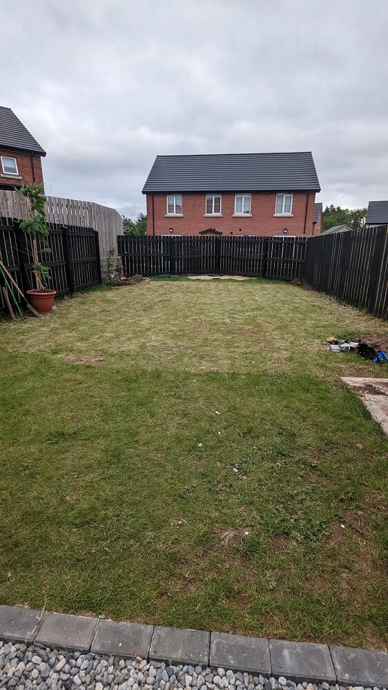 A spacious backyard with uneven grass, a flowerpot, and scattered garden tools against a black wooden fence.