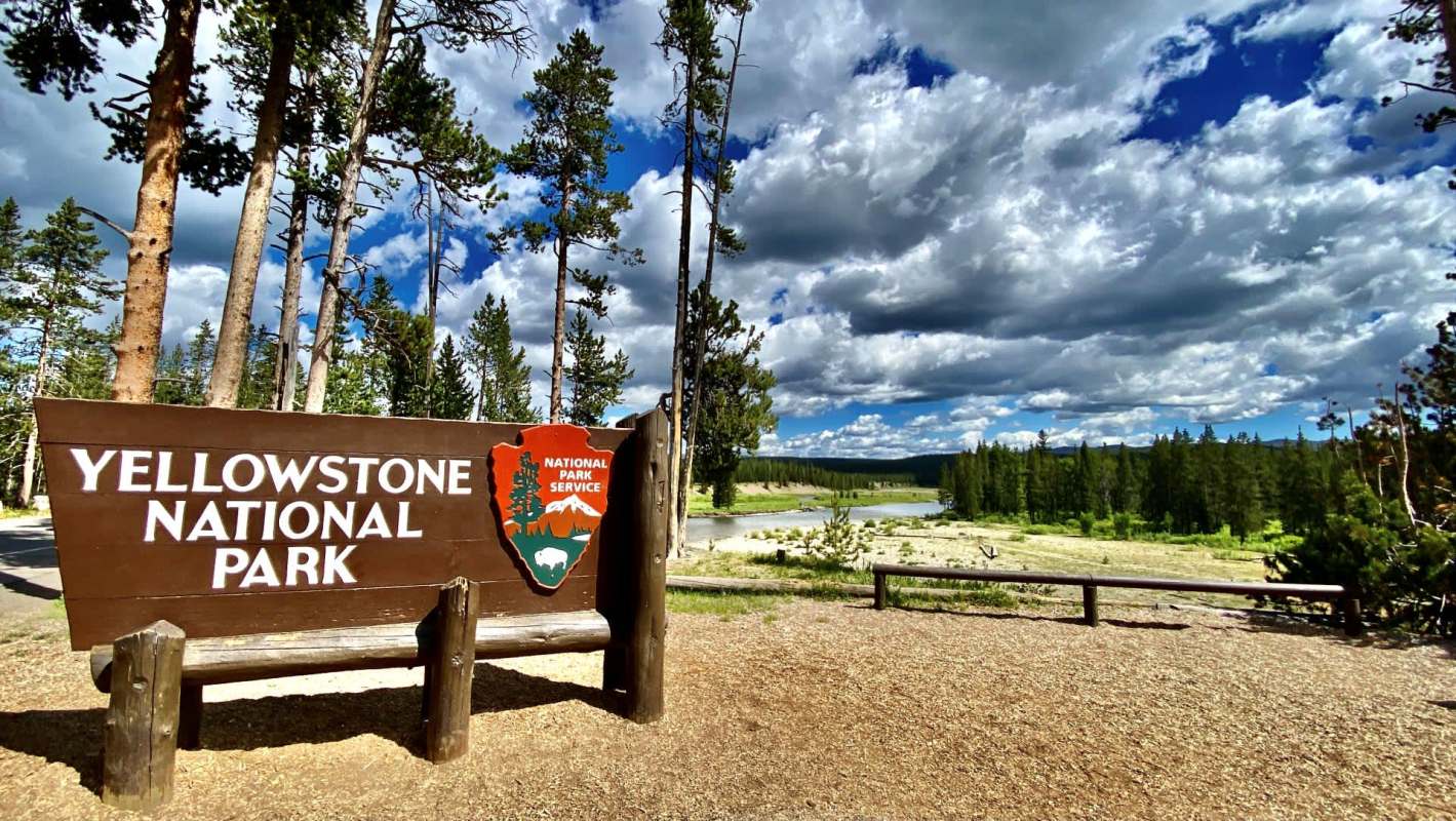 A wooden sign for Yellowstone National Park stands against a backdrop of trees and a cloudy sky.