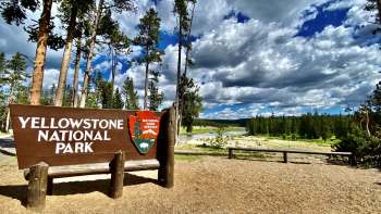 A wooden sign for Yellowstone National Park stands against a backdrop of trees and a cloudy sky.