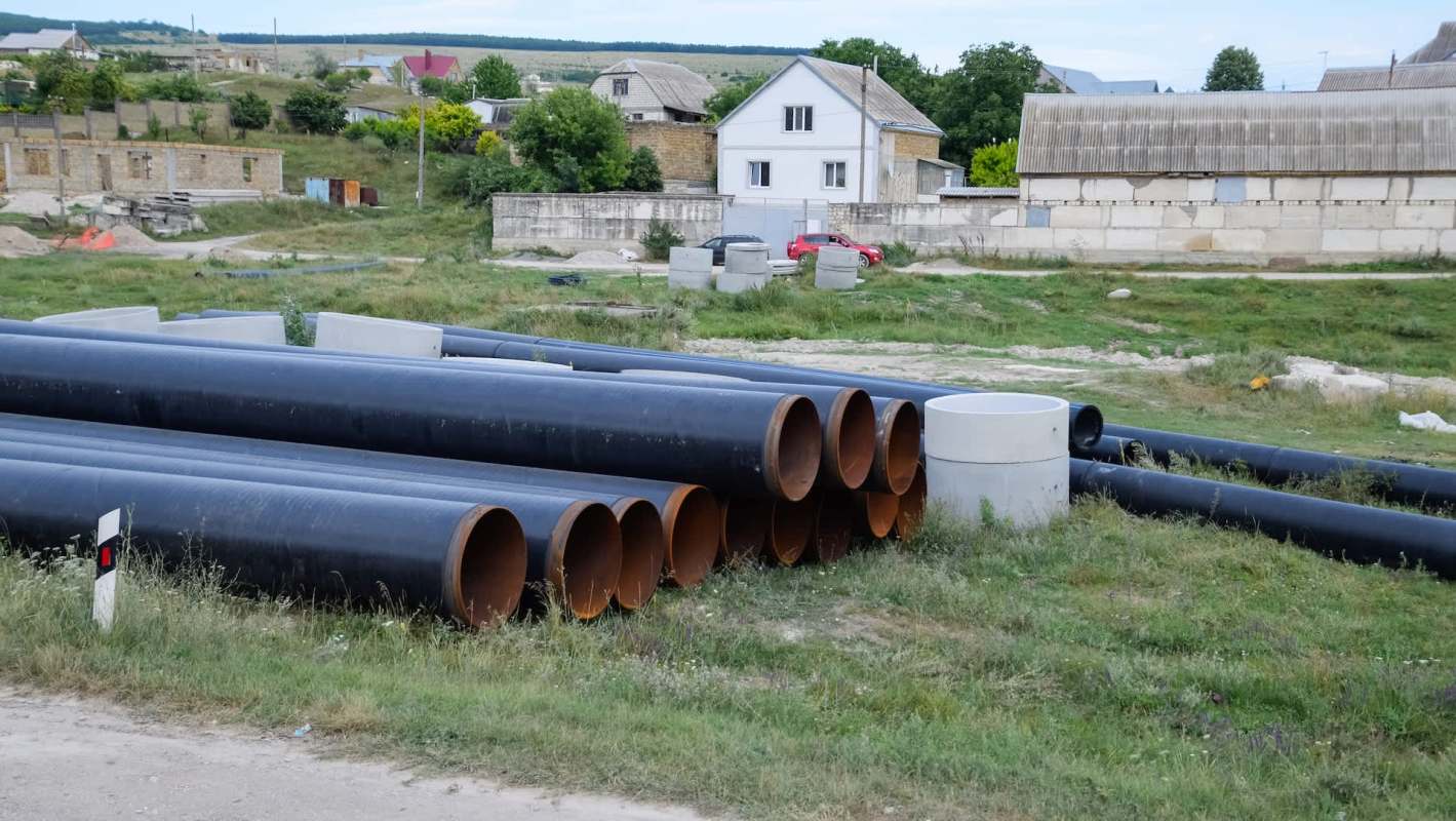 Piles of large black pipes and concrete structures are scattered in a grassy area near residential buildings.