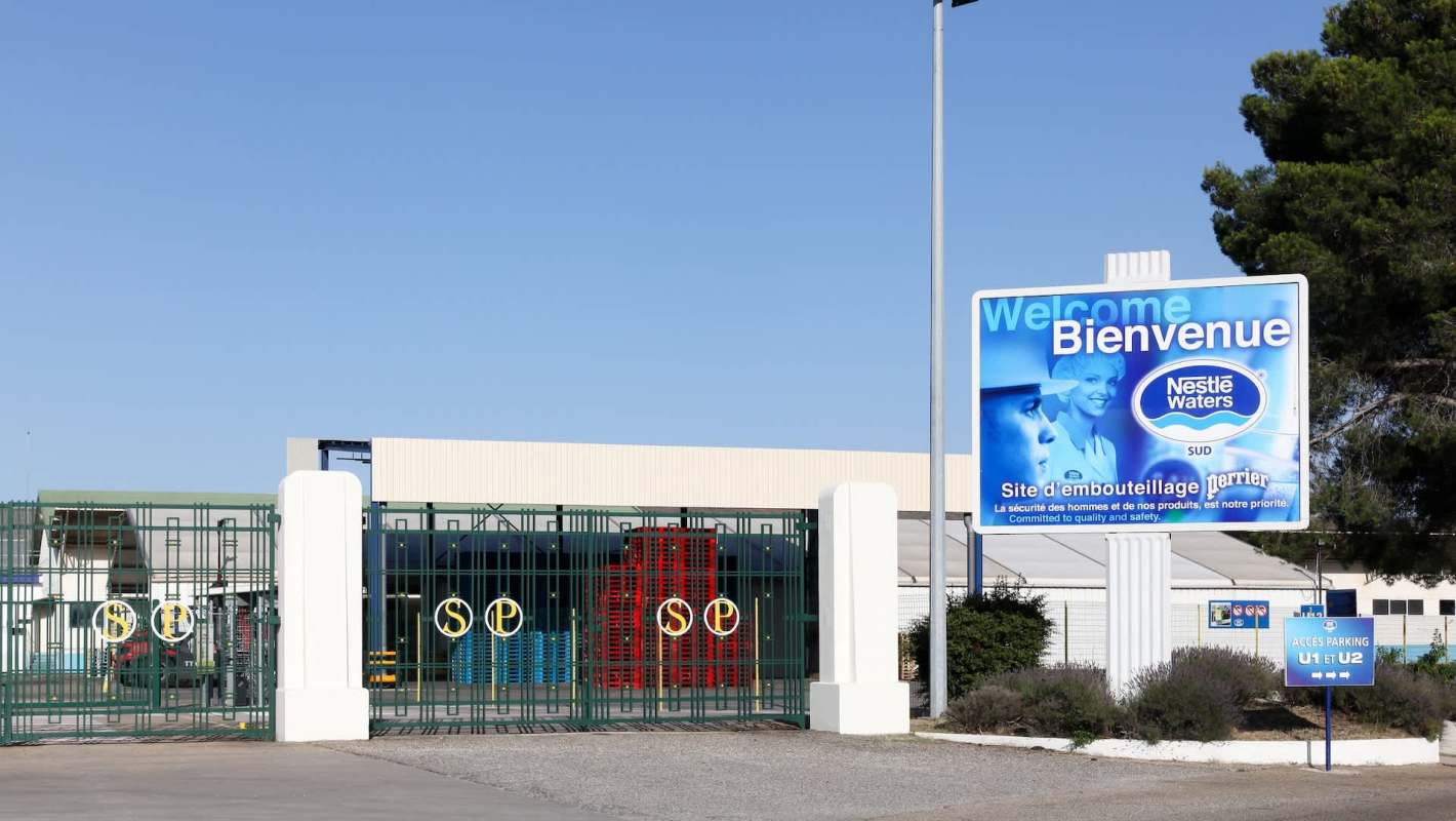 A factory entrance with a green gate, signage for Nestlé Waters, and colorful pallets in the background.
