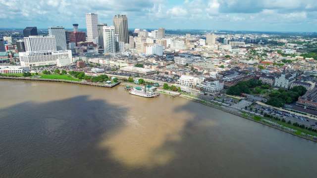 Aerial view of a city skyline along a river, featuring buildings and greenery near the water's edge.