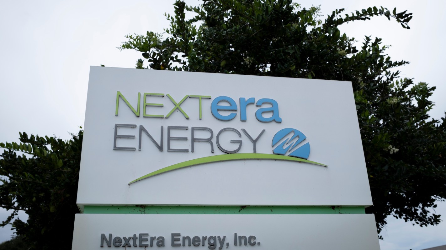 A close-up of the NextEra Energy sign displayed outdoors against a blurred green backdrop.