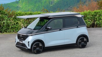 A compact modern electric vehicle with a roof-mounted solar system parked on a road with greenery in the background.