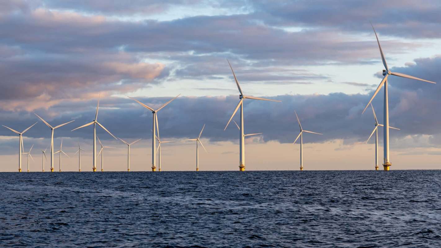 A row of wind turbines stands in the ocean under a cloudy sky at sunset.