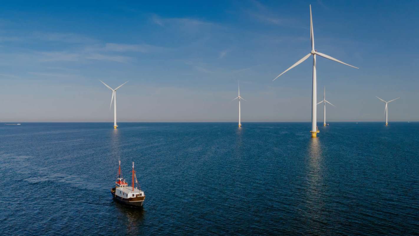 A boat sails near offshore wind turbines in a calm sea under a blue sky.