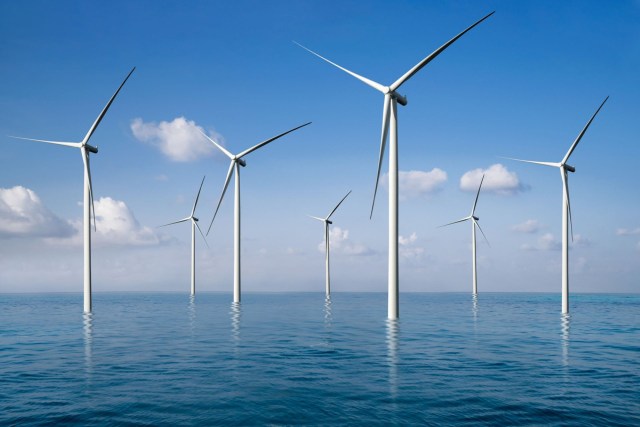 A row of wind turbines stands in a calm ocean under a clear blue sky with a few clouds.