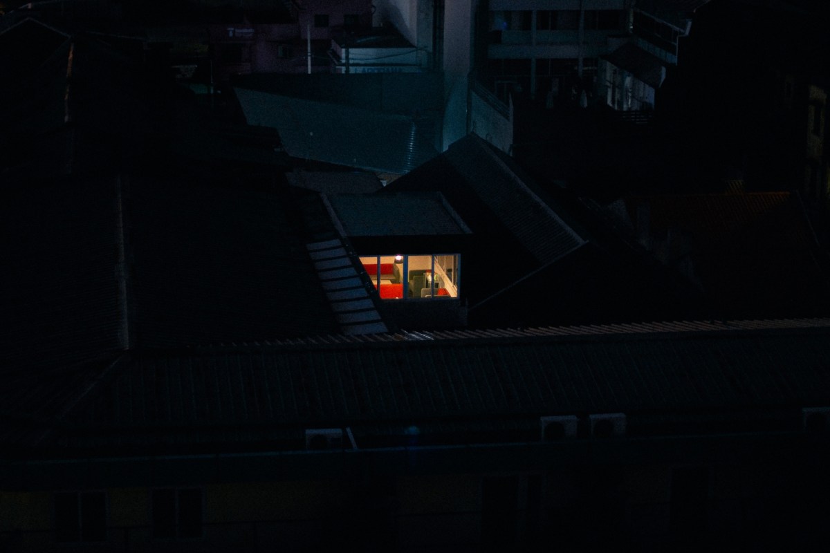 A brightly lit room in a dark urban setting surrounded by rooftops at night.