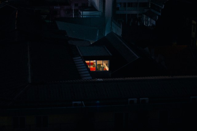 A brightly lit room in a dark urban setting surrounded by rooftops at night.