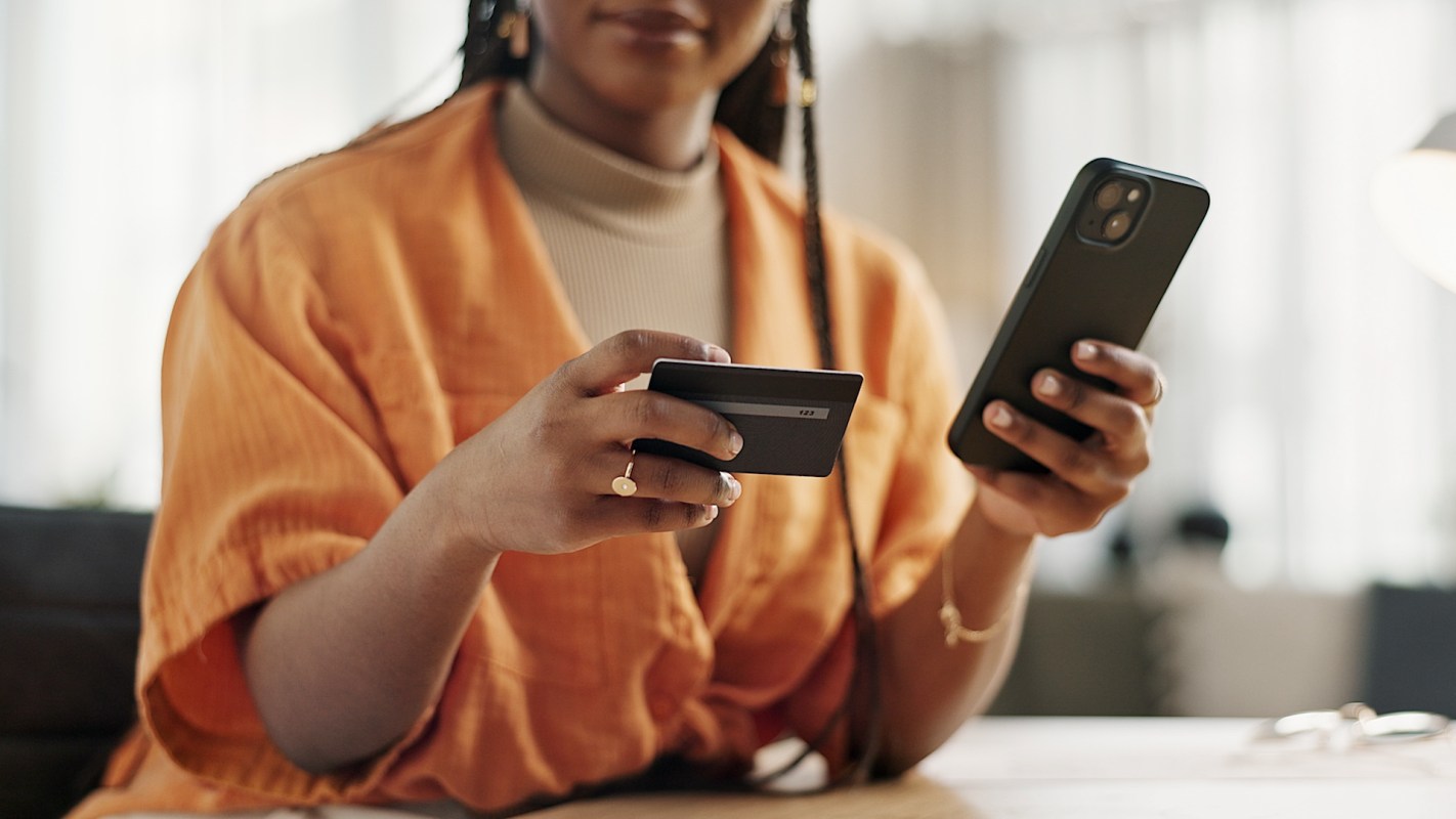 A woman in an orange shirt holds a phone in one hand and a credit card in the other while sitting at a table.