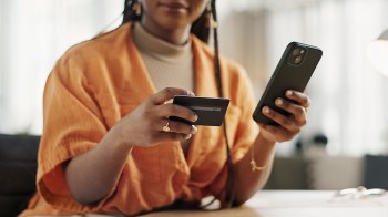 A woman in an orange shirt holds a phone in one hand and a credit card in the other while sitting at a table.