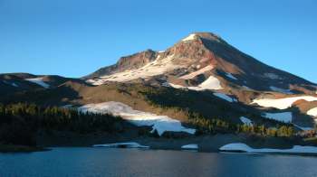 A towering mountain with snow patches and a clear blue sky, reflected in a calm lake in the foreground.
