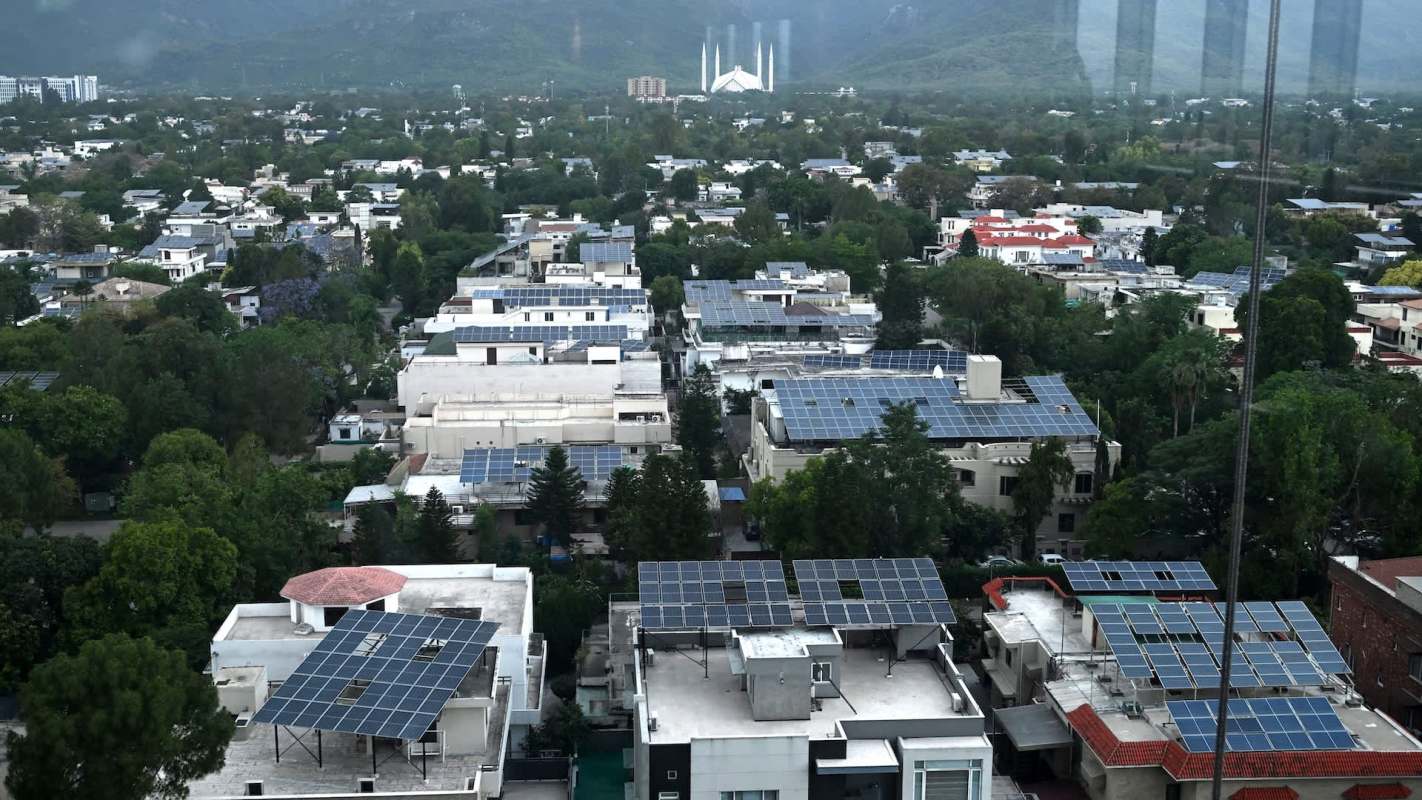 Aerial view of a neighborhood in Pakistan with homes featuring solar panels and lush green trees, against a mountainous backdrop.