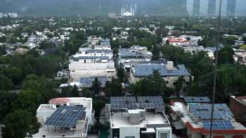 Aerial view of a neighborhood in Pakistan with homes featuring solar panels and lush green trees, against a mountainous backdrop.