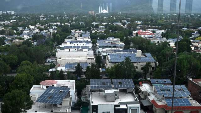 Aerial view of a neighborhood in Pakistan with homes featuring solar panels and lush green trees, against a mountainous backdrop.
