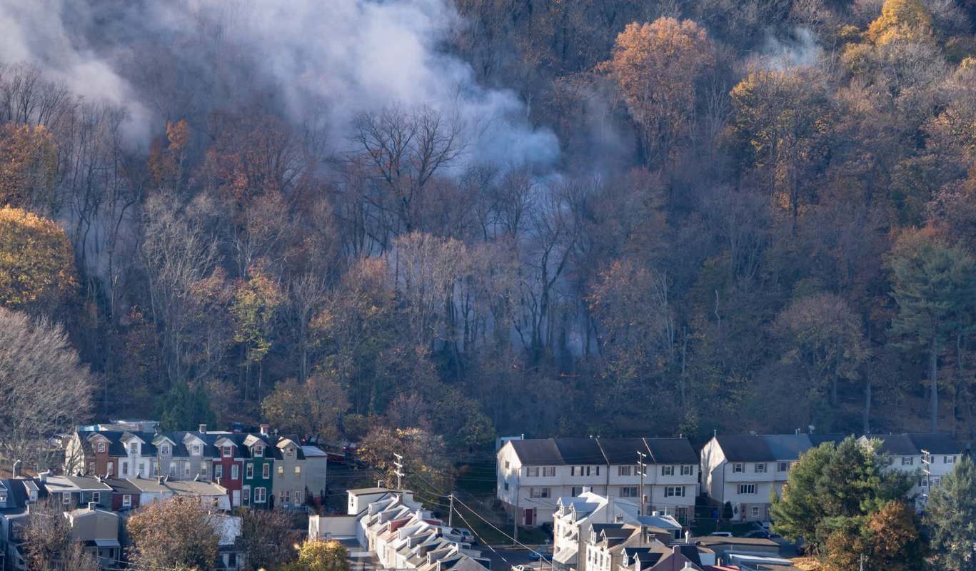 Wildfire smoke rises from a forested area behind a residential neighborhood with autumn foliage.