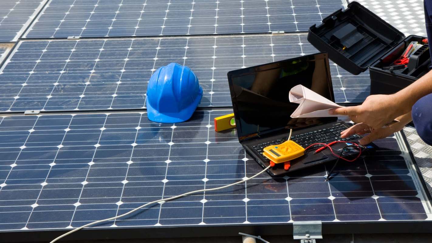 A worker analyzes solar panels using a laptop and testing equipment, with a blue hard hat nearby.