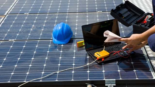 A worker analyzes solar panels using a laptop and testing equipment, with a blue hard hat nearby.