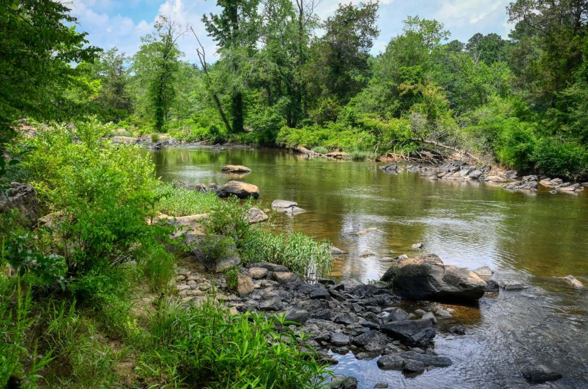 A serene river flows through a lush green landscape, bordered by rocks and trees under a partly cloudy sky.