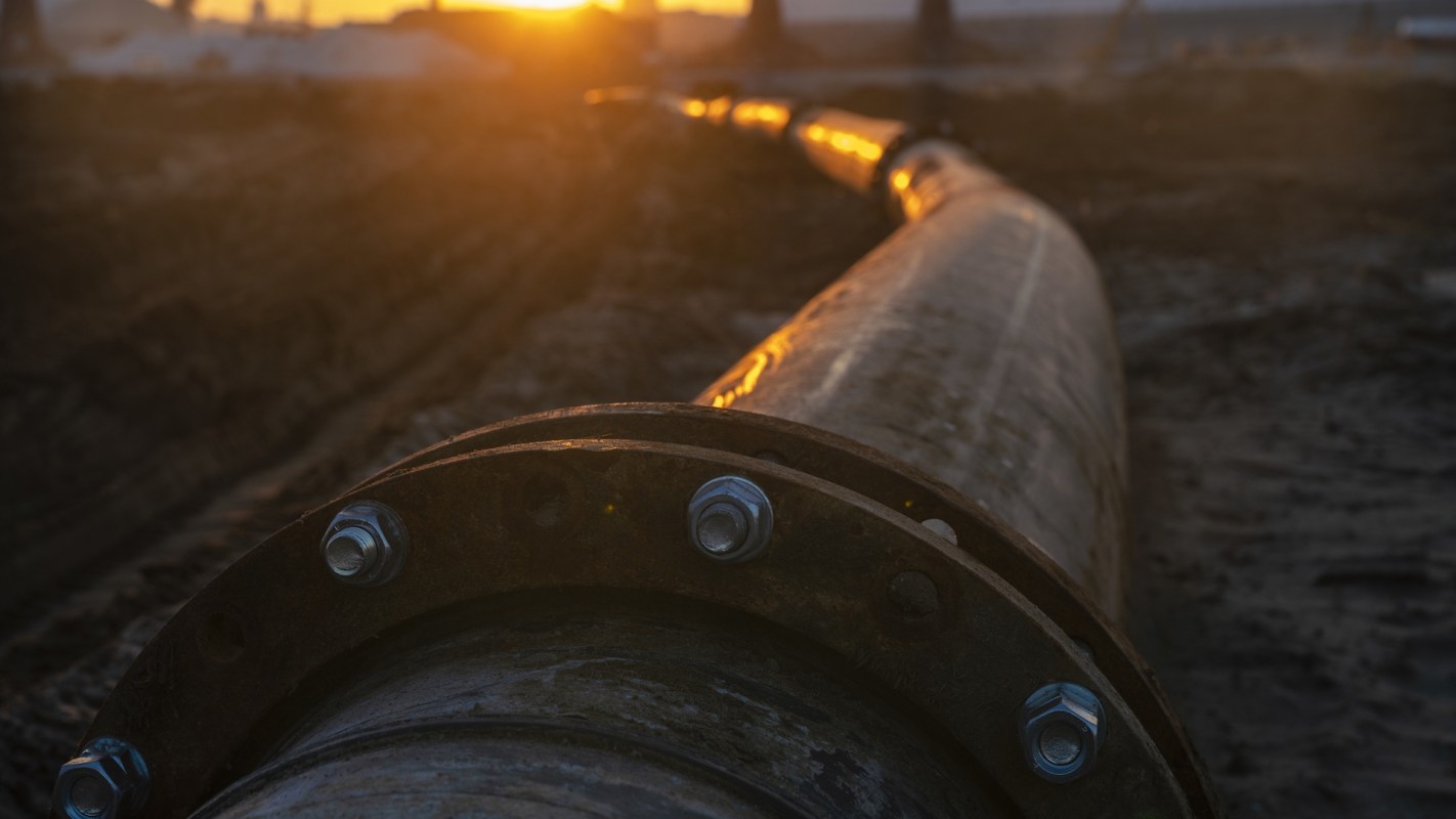 A close-up of a large metal pipe with bolts, illuminated by the warm glow of a sunset in the background.