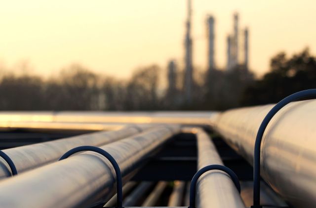 Industrial pipelines on a platform, with a refinery in the background.