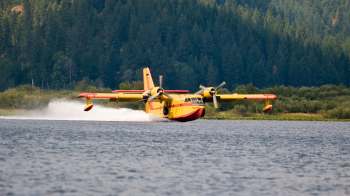 A yellow and red seaplane skims across a lake surrounded by trees.