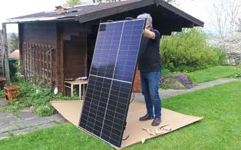 A person installs a solar panel in a garden beside a wooden shed.