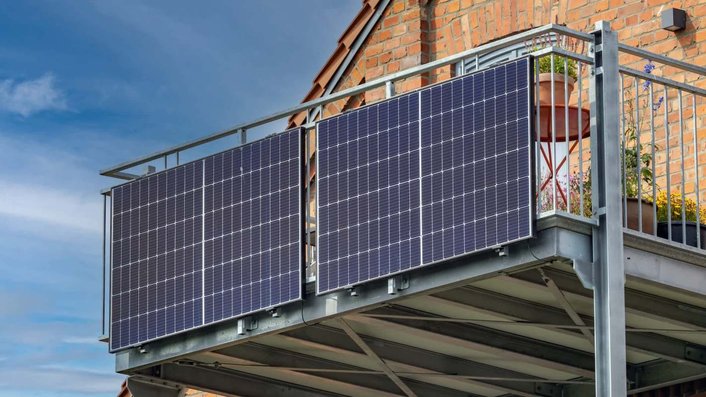 Two solar panels mounted on a balcony railing with a brick wall and potted plants in the background.