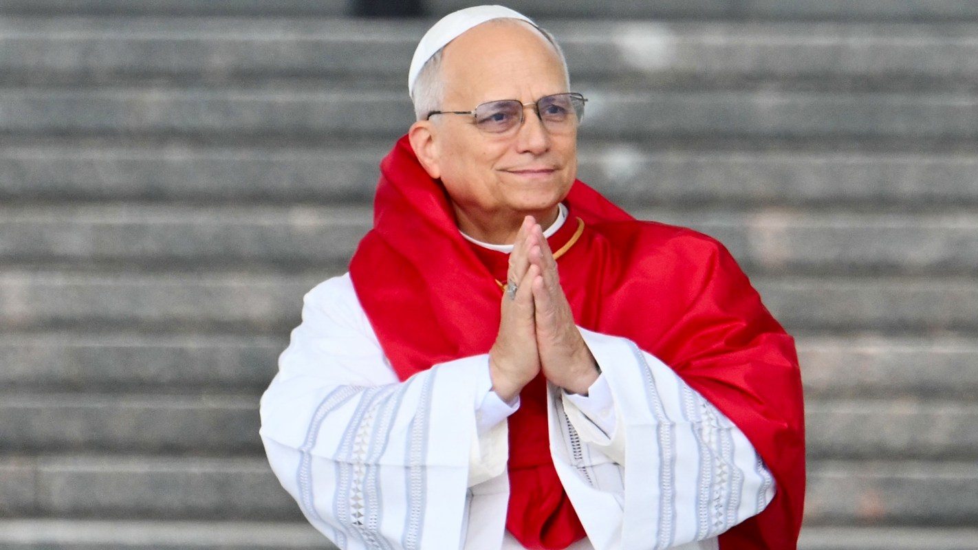Pope Leo XIV, wearing glasses and a red shawl, clasps his hands together in prayer.