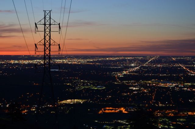 A silhouette of a power transmission tower against a sunset over a city illuminated by countless lights.