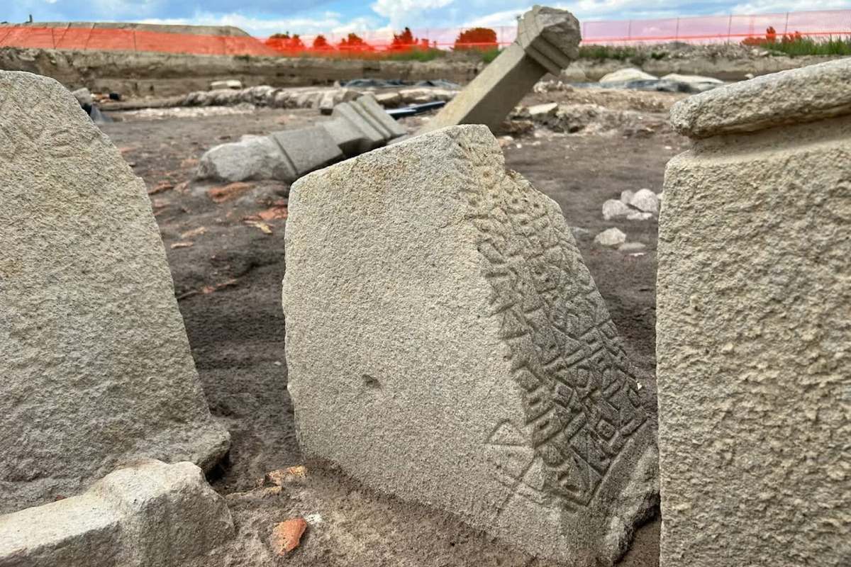 Ancient stone carvings are partially buried in sandy soil near construction barriers.