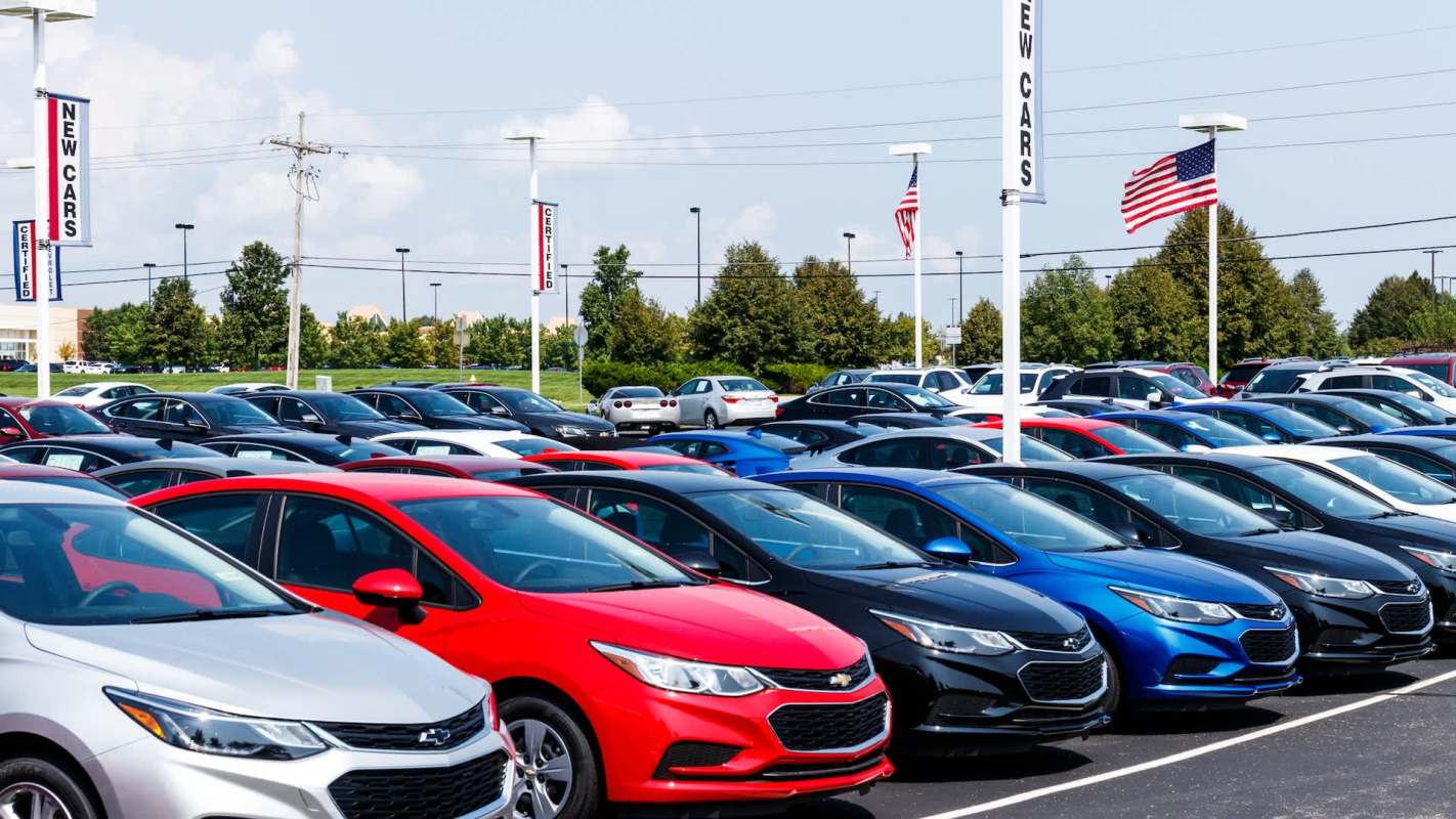A colorful assortment of new cars parked in a dealership lot with flags and signage in the background.