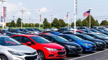 A colorful assortment of new cars parked in a dealership lot with flags and signage in the background.