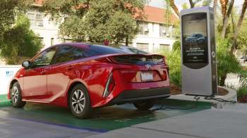 A red Toyota Prius plugged into an electric vehicle charging station surrounded by greenery.