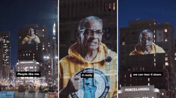 A projection of a elderly woman in a yellow hoodie speaking on a projection on a building at night.