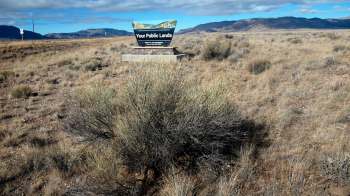 A sign reading "Your Public Lands" stands in a dry, open landscape with sparse vegetation and distant mountains.