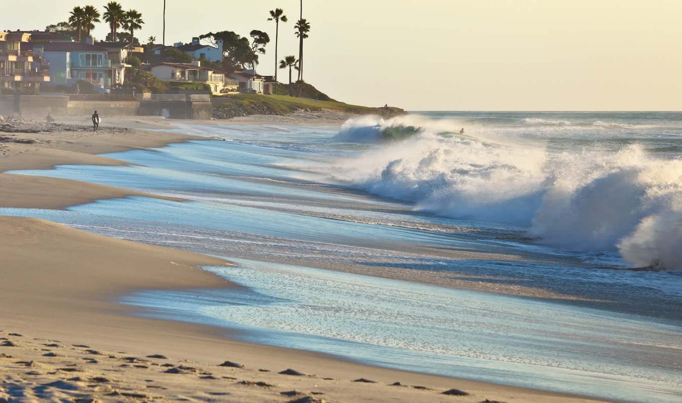 A person holding a surfboard on a beach with buildings and trees in the background during sunset and waves coming ashore.