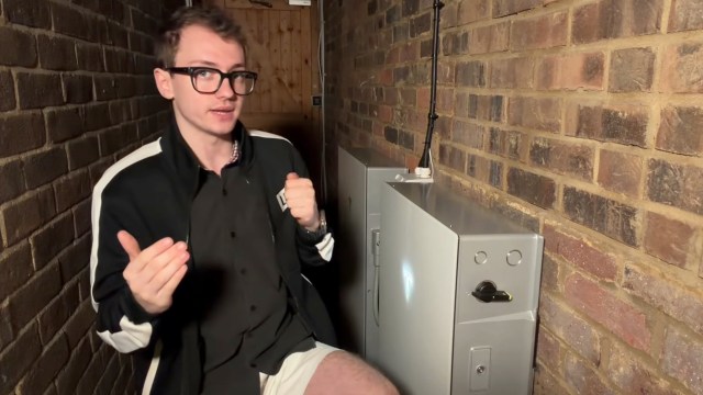A young man in a black jacket gestures while sitting next to a Tesla Powerwall with a metallic cover against a brick wall.