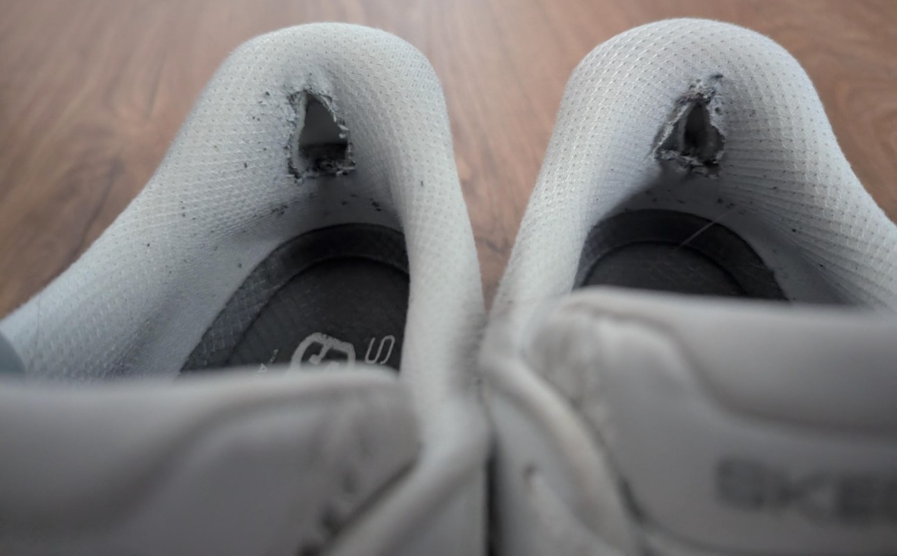 A close-up of worn-out white sneakers showing torn fabric at the heel area.